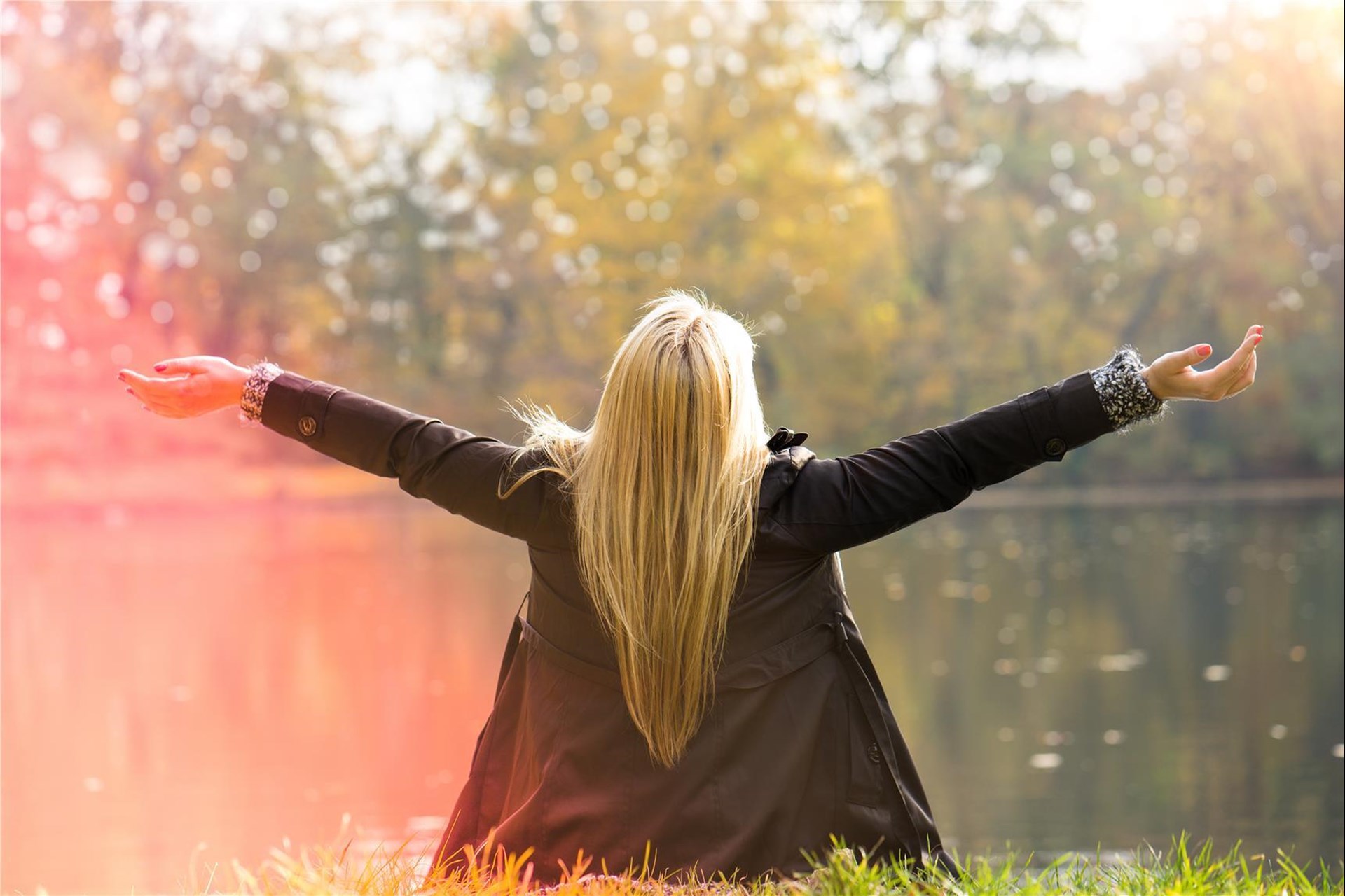 Blonde langhaarige Frau von hinten fotografiert sitzt mit ausgebreiteten Armen im Sonnenschein am Ufer eines Sees