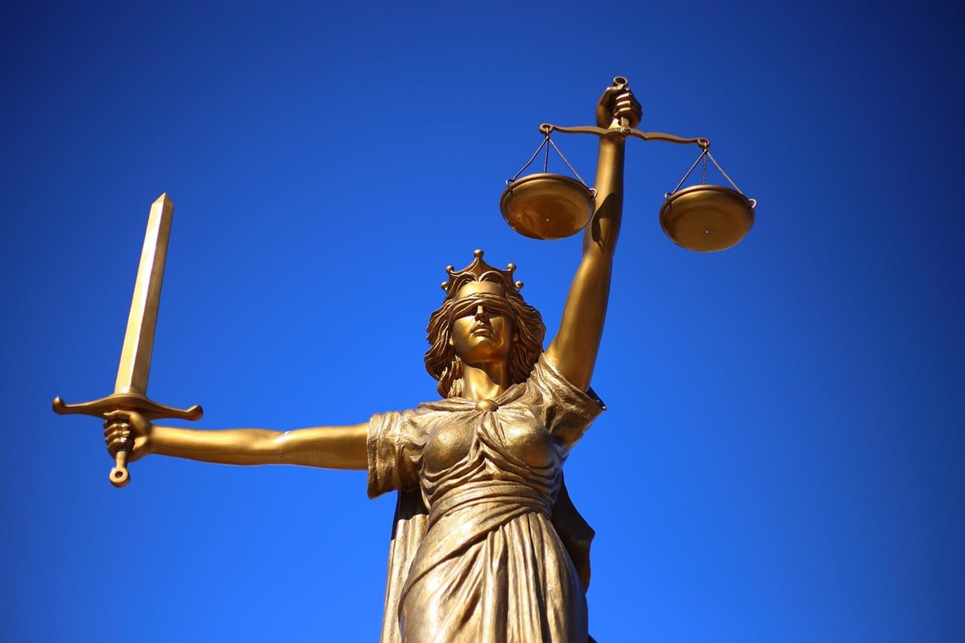 Bronzestatue der Göttin Justitia mit verbundenen Augen, Schwert und Waage vor tiefblauem Himmel