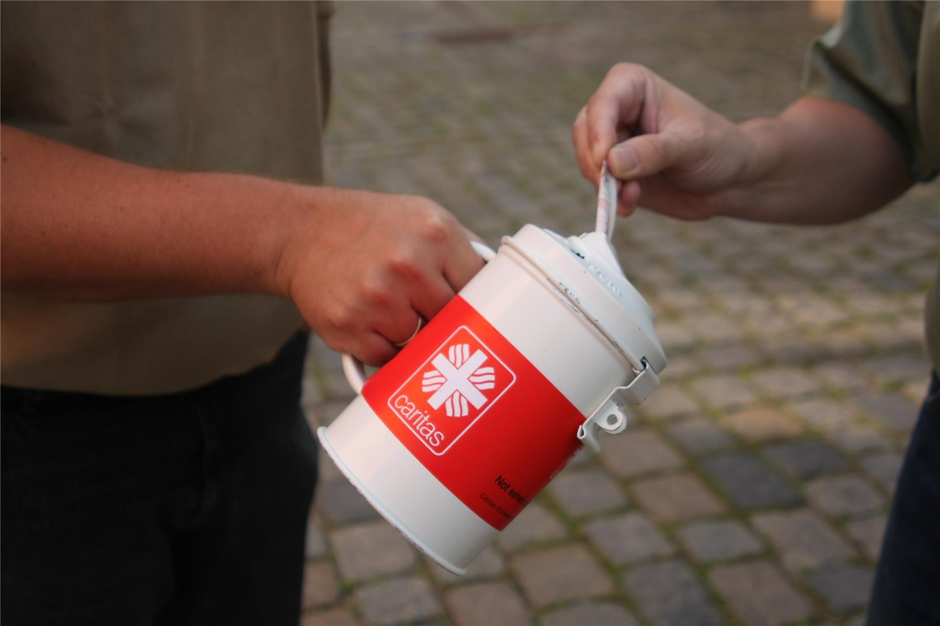 Bildausschnitt zweier Hände: Die eine hält am Henkel eine mit Caritas-Logo beschriftete Spendendose, die andere steckt einen Geldschein hinein
