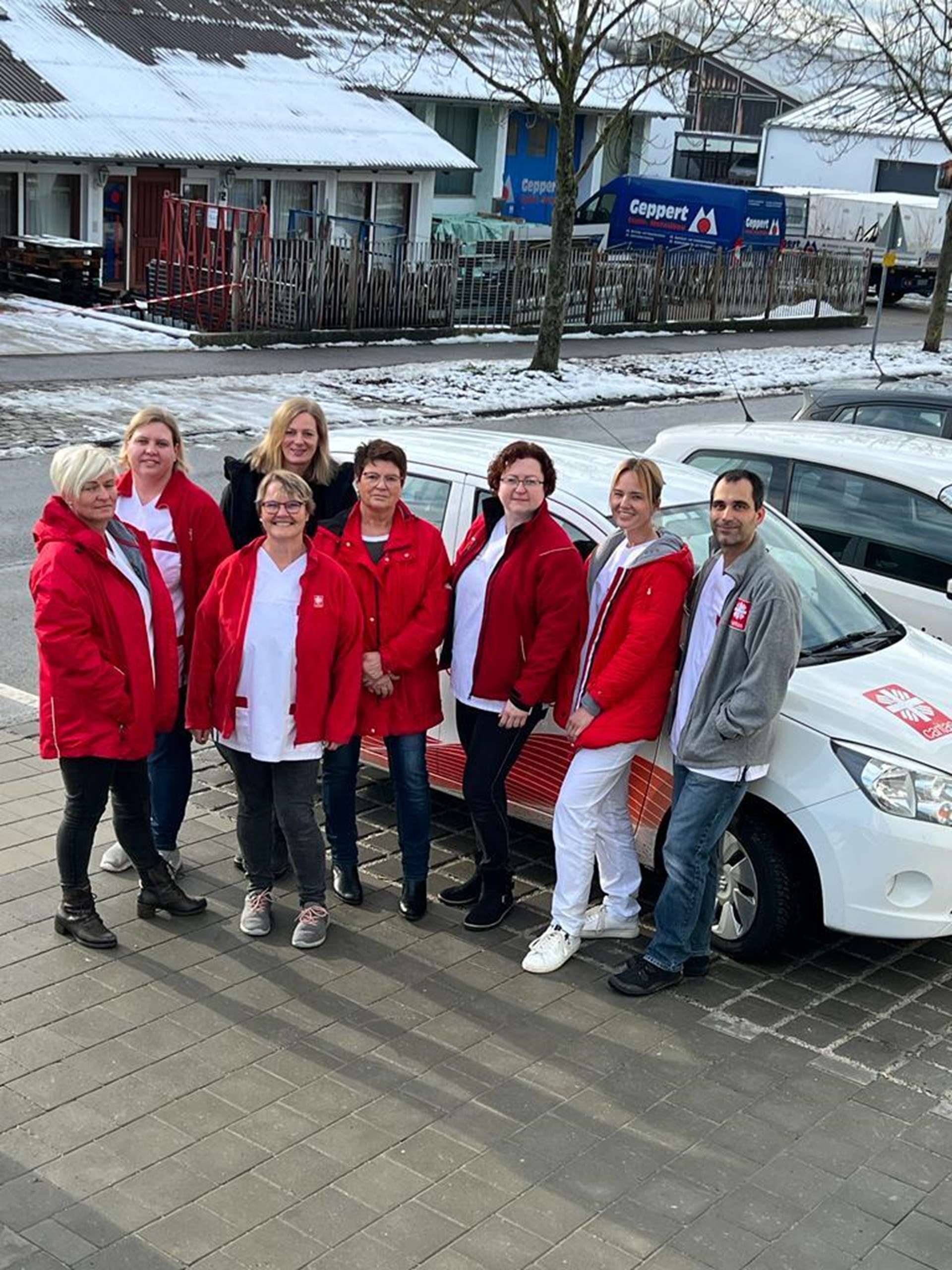 Das Bild zeigt das Team der Sozialstation Bad Abbach vor dem Dienst-Auto der Caritas. Die Pflegenden tragen die Dienstkleidung der Caritas und stehen vor dem Gebäude der Sozialstation.