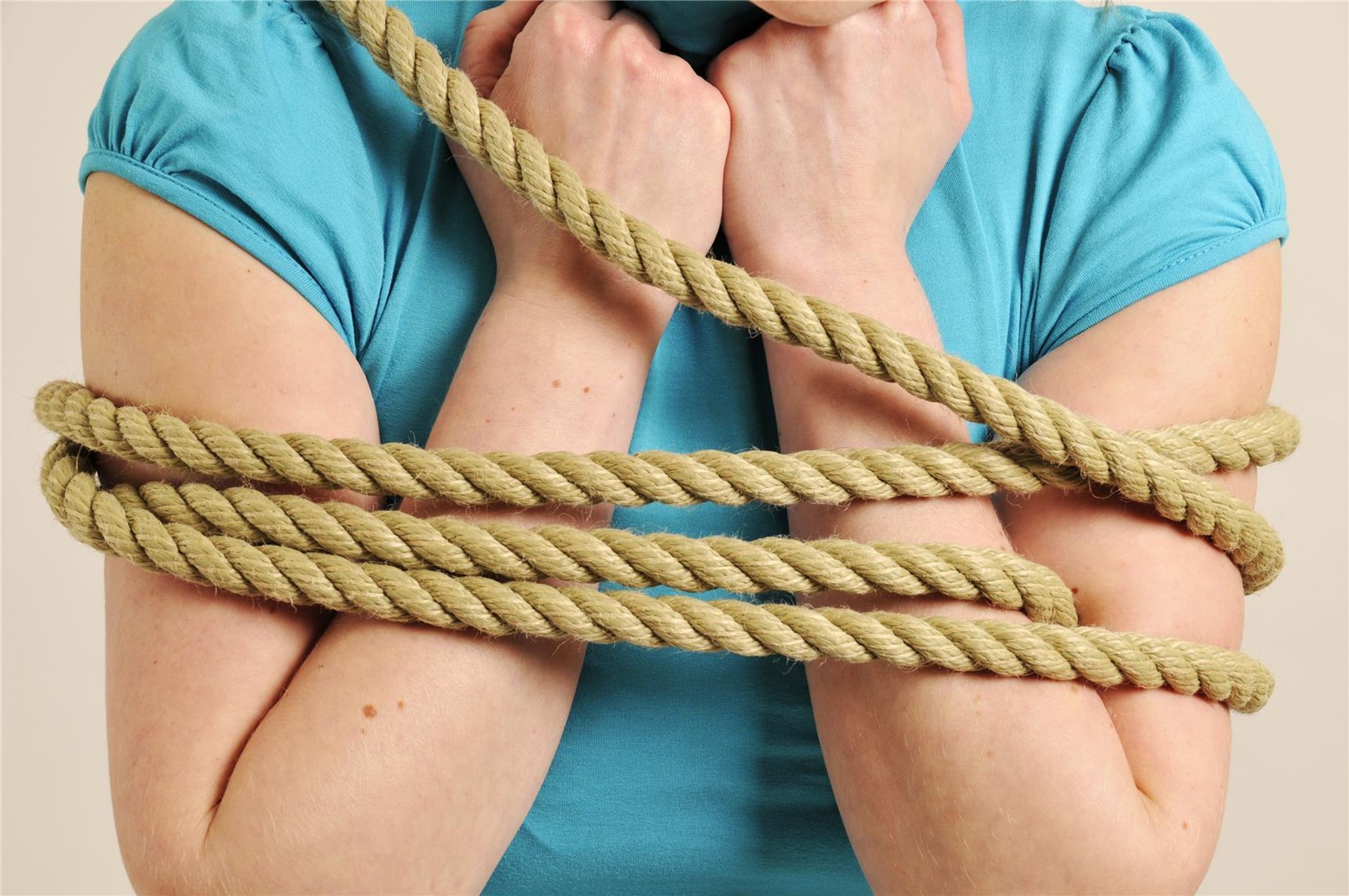 Bild vom Oberköper einer Frau, bekleidet in hellblauem T-Shirt., die Arme vor der Brust verschränkt mit den Händen unterm Kinn. Die Arme sind mit einem dicken Hanftau gefesselt